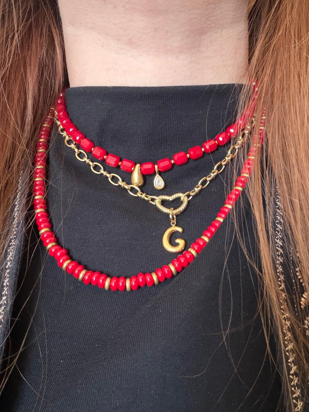 Drie gelaagde handgemaakte rode kralenkettinkjes met gouden bedels en letterbedel G, gedragen op zwart shirt